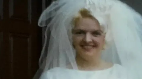 Beryl Davies, who has blonde hair and a white wedding veil, smiles at the camera