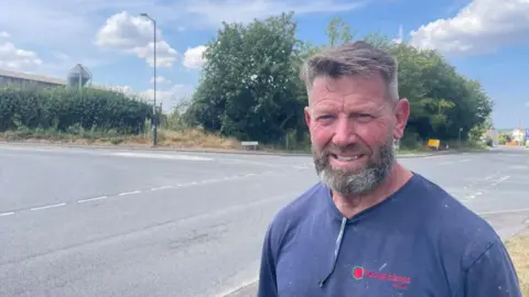 A man is standing to the right of some crossroads, smiling at the camera. He has brown hair and a greying beard and is wearing a blue t-shirt with sunglasses tucked into the neck.