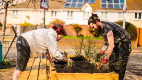 Two people facing opposite each other. They are leaning over a wooden plant pot. There are both planting plants. Behind them are houses and a road. 
