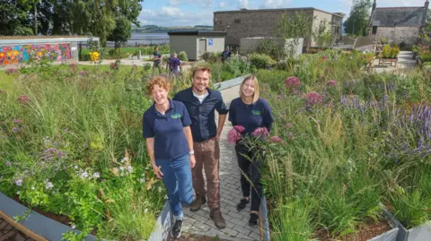 Three people - two women flanking one man - standing in the middle of a Saltire shaped garden. They are all laughing and smiling while surrounded by greenery.