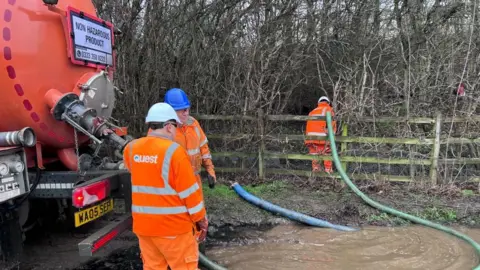 Three men in orange high visibility suits position hoses into muddy water in a woodland.