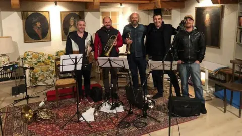 Facebook Five men standing in a recording studio, three of whom are holding brass instruments. Stands with music sheets are set up in front of them.