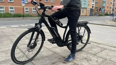 BBC A generic photo of a person on a pavement astride a black e-bike, showing their legs, one of which is on a pedal. They are wearing dark trousers and blue trainers and there are red brick flats and a road in the background