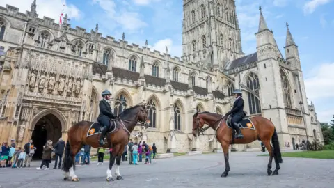 Two police officers straddled on horses outside of Gloucester Cathedral on a sunny day