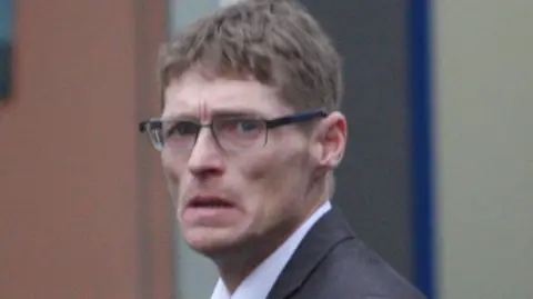 Central Scotland News A skinny man with brown hair and glasses. He looks worried and is wearing a suit. 