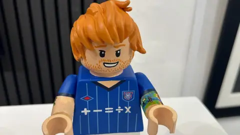 A general view of the Ed Sheeran Lego figure. It shows the figure with ginger hair and a smile painted on his face with ginger stubble. It is painted in a blue Ipswich Town football shirt and the arms have been raised. The right arm has a tattoo painted on.