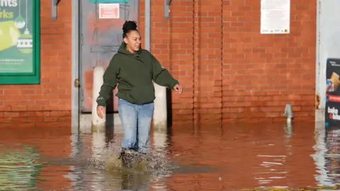 PA Media A woman dressed in a hoodie and jeans walks through floodwater outside a building at the Billing Aquadrome in Northamptonshire.