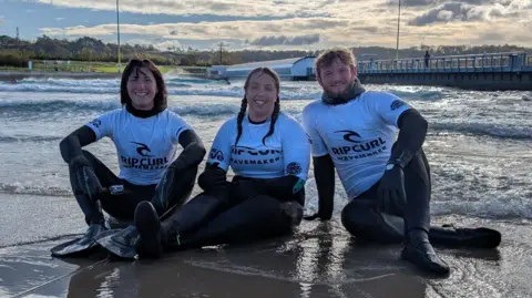 Three people sit on the shore of an artificial beach in Bristol. In the middle is Kay Millar, who is sitting with her legs outstretched in front of her. Her hair is tied in tow ponytails. On her right is her coach Hannah Mattison, who has shoulder-length brown hair. On Kay's left is her coach Ryan Trott who has short brown hair. They are all wearing black wetsuits with white Rip-Curl rash vests on top.