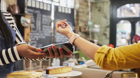 Tara Moore/Getty Images Creative Young woman paying for goods with a smart watch in a coffee shop