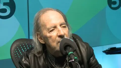 Harry Shearer has long grey hair and is wearing a black leather jacket. He is sitting in front of a microphone.