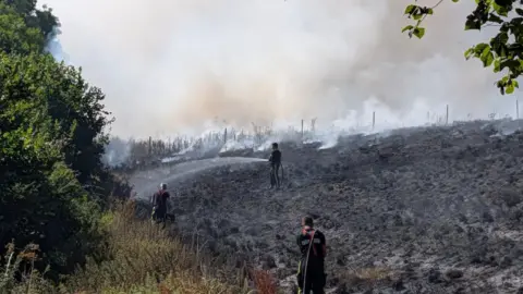 Devon & Somerset Fire & Rescue Service Three Devon & Somerset Fire & Rescue Service firefighters tackling a large wildfire in a field which is completely burnt and black following the fire.