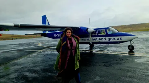 Tammy Hine Tammy Hine, standing in front of a plane, that is blue and white in colour. She has on a long coat, a pink cardigan, blue tights, a dress and a scarf. She is standing on tarmac and fields are behind her. 