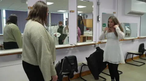 A young girl models for a white dress in front of a mirror with the help of an older woman in a cradigan