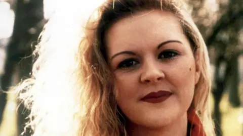 A close-up photo of Fiona Pender, a woman with blonde curly hair, looking directly at the camera. She is wearing red lipstick and a red top.
