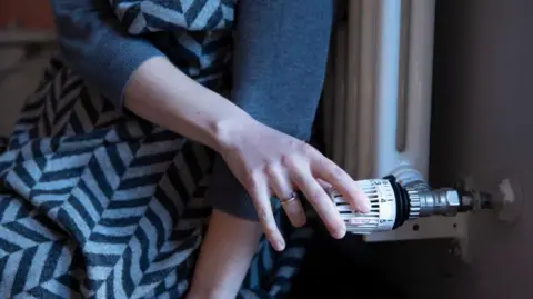 Getty Images A close-up of someone with their back to a radiator and a blanket over them, turning to adjust the temperature on the dial.