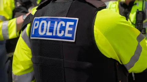 This picture shows three police officers in uniform. None of the officers are identifiable. They are wearing a high-vis jacket with a black short-sleeved jacket. On the back of one of their jackets it says Police 
