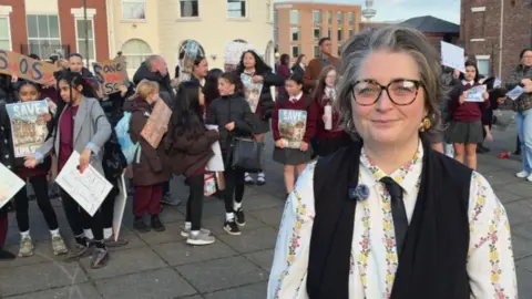 Katie Brennan has short grey hair and is wearing dark framed glasses, a floral shirt, a black waistcoat and a black tie. She is standing in front of a group of children who are holding banners which read "Save Our School".