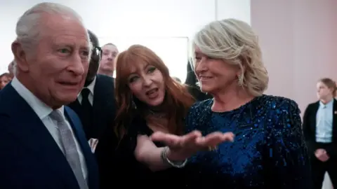 Reuters Makeup artist gestures as she stands between King Charles, in a blue suit, and Martha Stewart in a gliterring navy blue dress. 