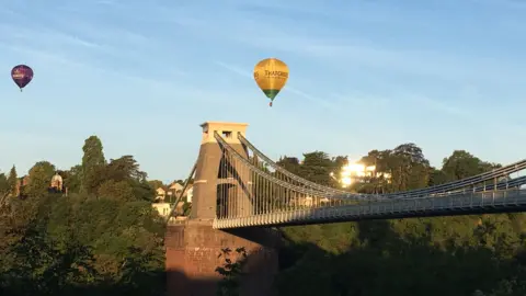 The Clifton Suspension Bridge with two balloons above it, one is yellow and sponsored by Thatchers cider and another is purple but with an unclear sponsor.