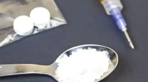 A spoon with a white drug in on sits next to a bag of drugs and an injection on a dark table. This is a stock shot. 