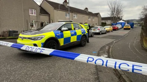 A uniformed police officer stands behind a police car on a street which has been cordoned off with police tape. There are several cars parked outside houses and police forensic tents can be seen in the background