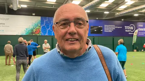 BBC A mid-shot of John Adams. He has grey hair, wears glasses, and has a blue jumper. In the background are the carers and others playing sport.