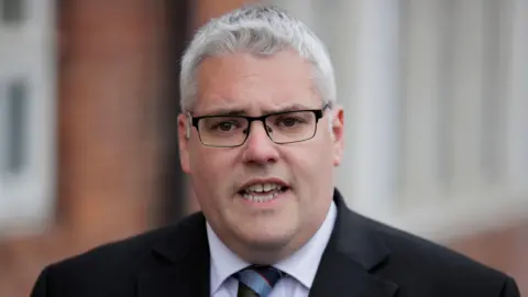 PA Media Gavin Robinson, who is wearing black-rimmed rectangular glasses and a black blazer and white shirt, has is mouth open, showing both rows of teeth, as if in the middle of speaking. His grey fringe rises in a cow's lick and his face is broad and full. 
