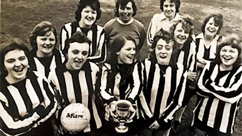 A black and white image of a 11 women in football uniforms with black and white vertical stripes. They are all smiling. In the centre of the group one of the women is holding a trophy, while the woman next to her is holding a football