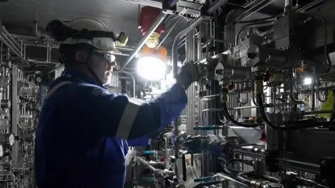 BBC A man in a blue boiler suit wearing a hard hat stands in front of machinery.