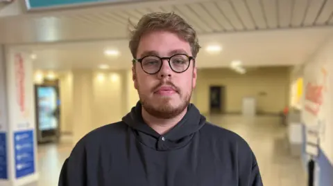 James Bayliss-de Gruchy is looking steadily at the camera as he stands in the airport. He has short wavy brown hair and a beard. He is wearing glasses and a black hoody.