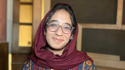 Hermain is wearing a patterned outfit with a maroon head covering stands indoors in front of a wooden structure with rectangular sections. The background includes warm-toned walls and dim lighting.