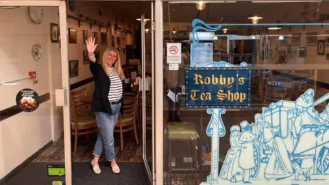 Ruth Carvell Doyle A woman with blonde hair who is standing in the doorway of a shop waving goodbye. She is wearing jeans and a stripy top and the shop has a sign "Robby's Tea Shop" in the window