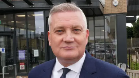 John Fairhall/BBC Russell Quirk is wearing a navy suit jacket, white shirt and dark tie. He has short grey hair and is standing in front of a building with a glass facade.