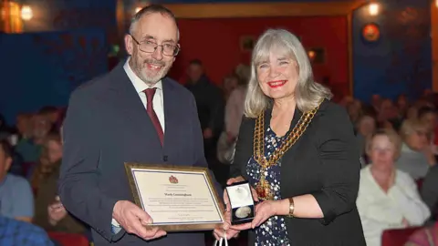 The cinema owner Mark Cunningham, dressed in a smart dark grey suit, white shirt and red tie, is holding an A4 sized, gold framed certificate bearing his name and the crest of Gloucester City Council. Next to him is the Mayor, Lorraine Campbell, wearing her ceremonial chain on top of her dark jacket and blue flowery dress. She is holding a small, open presentation box containing a silver circular medal. They are standing inside the cinema, with a small audience in their seats behind them.