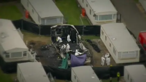 Aerial shot of caravan park, showing eight white caravans, with the centre one a burnt out shell. Nine white suited figures can be seen examining the charred interior and immediate area outside