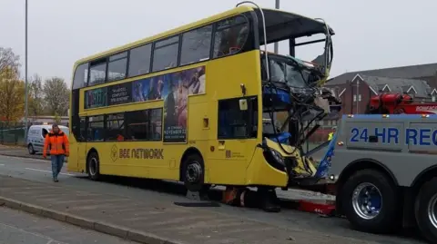 A yellow Bee Network bus being towed away by a recovery vehicle as a man in orange high-vis jacket watches on.