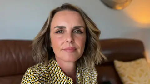 BBC news Rachel Williams looks into the camera. She is wearing a yellow, black and white blouse, and is sitting on a brown leather sofa.  