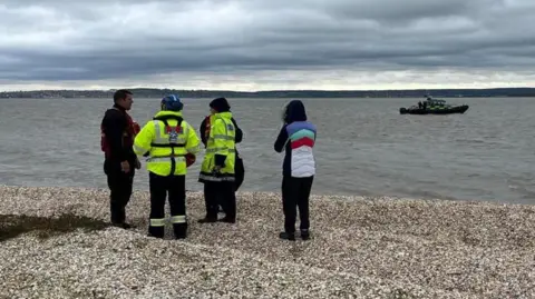 Chrissie Reidy/BBC Two people in high-vis coats with two others standing next to the shoreline with a boat in the river