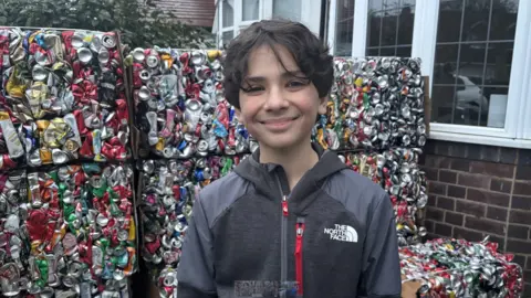 A boy with black hair and a grey coat stood in front of crushed aluminium cans