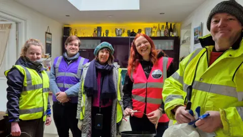 A group of five people - three women and two men - wearing high vis vests and carrying buckets. They are standing indoors in a kitchen.