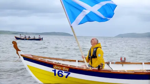 A man with a Scotland flag in a brightly coloured yellow, white and blue rowing boat called the Lady Bay