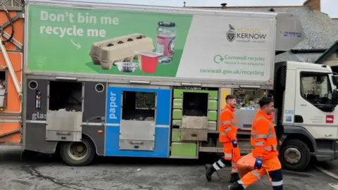 BBC Two waste disposal staff wearing high viz jackets, walking in front of a Cornwall Council recycling lorry.