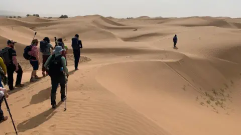 Alex Baines A group of people trekking across a sand dune in the Sahara desert.