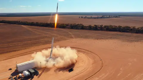 HyImpulse Technologies GmbH A rocket being launched from a brown, dusty landscape. There is fire coming out of the rocket itself and fields in the background. 