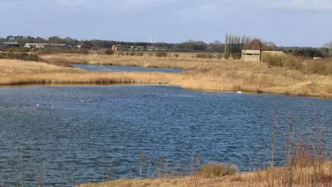 Pauline E / Geograph A view of one of the lakes at North Cave Wetlands the blue waters are surrounded by reeds and a wooden bird watching hide is on the opposite bank