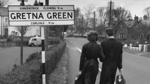 Getty Images A black and white photograph of a couple walking down a street with their back to the camera. A road sign on the left reads GRETNA GREEN in large text. Houses, power lines and hedges line the street.