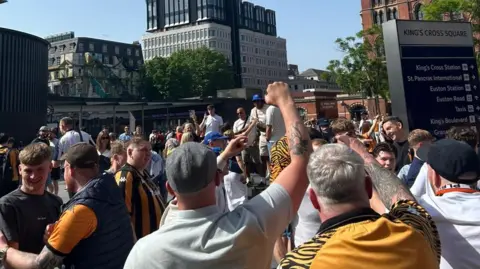 Submitted Hull City supporters, wearing black and amber shirts, cheer outside King's Cross station in London 
