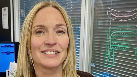 Close-up portrait of Emily Cutts with shoulder-length blond hair in an office setting, standing in front of window blinds decorated with colourful marker drawings. A computer monitor and office materials are visible in the background.