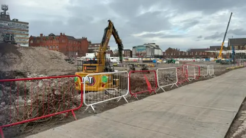 A yellow digger is on a construction site behind red and white fencing and a concrete path.