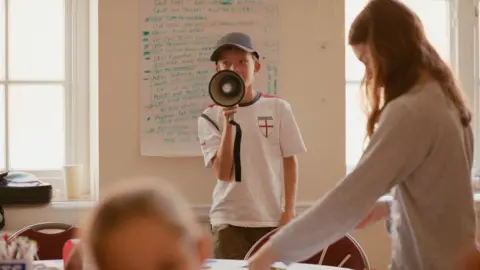 Boy in an England football shirt and a blue baseball cap, using a megaphone in a classroom